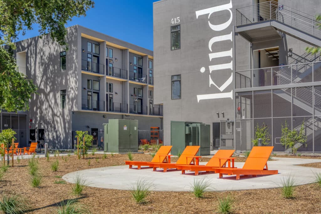 Four bright orange lounge chairs are arranged on a circular concrete area in front of a modern gray apartment building with balconies and large “kind” lettering on the wall.